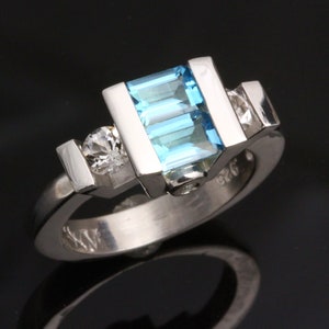 May include: A silver ring featuring a large, rectangular, light blue gemstone and two smaller, round, clear gemstones.