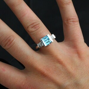 May include: A silver ring featuring a blue topaz gemstone and two smaller diamonds. The ring is worn on a finger.