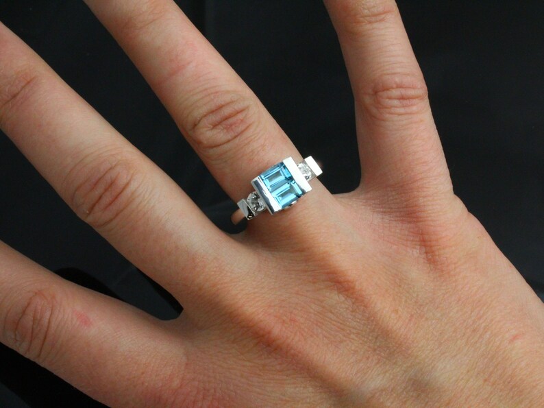 May include: A silver ring featuring a blue topaz gemstone and two smaller diamonds. The ring is worn on a finger.