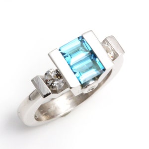 May include: A silver ring featuring a blue topaz gemstone and two smaller diamonds. The ring has a contemporary, geometric design.