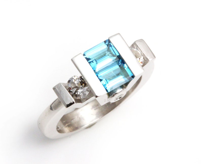 May include: A silver ring featuring a blue topaz gemstone and two smaller diamonds. The ring has a contemporary, geometric design.