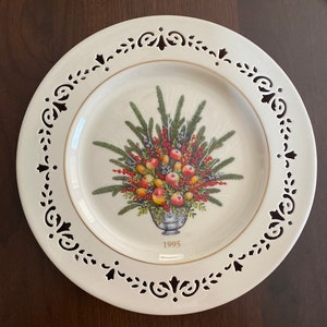 May include: A white ceramic plate with a gold rim and a black and white decorative border. The center of the plate features a colorful illustration of a floral arrangement with fruit and berries in a vase. The year 1995 is printed in gold at the bottom of the plate.