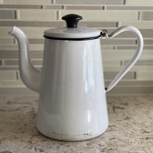 May include: White enamel coffee pot with a black lid and handle. The pot has a slightly worn look, with some scratches on the enamel.