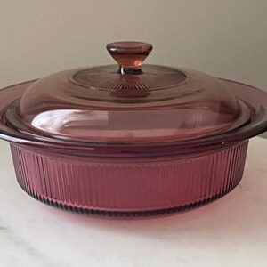 May include: A vintage, round, purple glass casserole dish with a matching lid. The dish has a ribbed exterior and a handle on the lid. The glass is a translucent, deep purple color. The casserole dish is likely used for baking and serving.