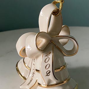 May include: Ivory-colored bell-shaped Christmas ornament with gold trim and a gold hanging cord. The ornament has a bow design and the year "2002" is printed in gold. The words "our Christmas" are also printed on the ornament.