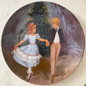 May include: A vintage porcelain plate with a painting of a young couple dancing in front of a Christmas tree. The girl is wearing a white dress with a red sash and the boy is wearing a black suit and white shirt. The plate is signed "Shell Zver" in the bottom right corner.