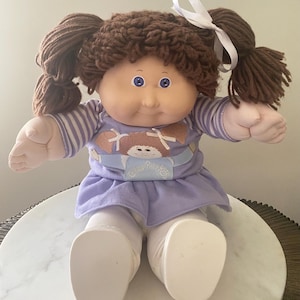 May include: A Cabbage Patch Kids doll with brown yarn hair in pigtails, wearing a purple and white striped shirt with a Cabbage Patch Kids logo, a purple dress, and white pants.