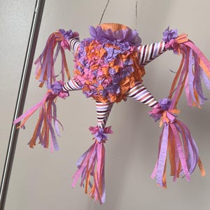 May include: A colorful star-shaped piñata with orange, pink, and purple crepe paper decorations. The piñata has striped arms and long, flowing streamers in shades of pink, orange, and purple. It is suspended from a silver wire.