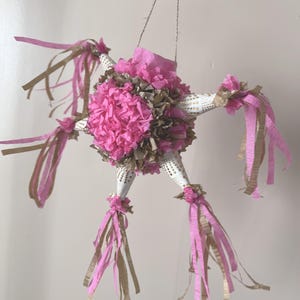 May include: A handmade star-shaped piñata with pink and gold streamers. The piñata is decorated with pink and brown paper flowers and has a white and gold patterned body. It is suspended by a twine string.
