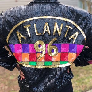 May include: A black sequined jacket with a large gold and colorful patch on the back that says "ATLANTA 96".