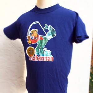 May include: A navy blue t-shirt with a cartoon illustration of a fisherman holding a fishing rod and a fish. The text "World's Greatest Fisherman" is in a gold circle and the text "Fisherman" is in red and white letters.