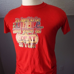 May include: Red t-shirt with a vintage-style graphic that reads "It's hard to be HUMBLE when you're as GREAT as I am!"