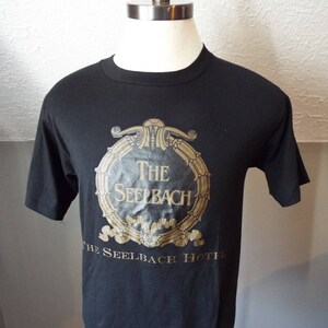 May include: Black t-shirt with a gold-colored circular design featuring the text "The Seelbach" and "The Seelbach Hotel". The design is centered on the front of the shirt.