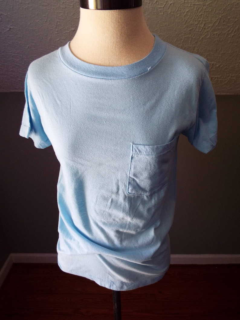 May include: A light blue t-shirt with a pocket on the left side.