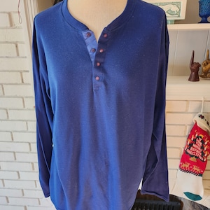 Può includere: Una camicia a maniche lunghe di colore blu scuro con colletto abbottonato. La camicia ha un'unica patta con bottoni ed è realizzata in un tessuto morbido e testurizzato.