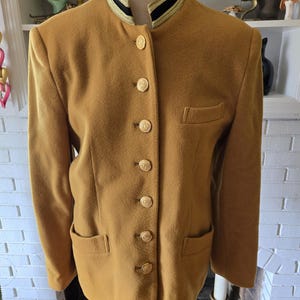 May include: A mustard yellow blazer with a black and gold trim collar and gold buttons. The blazer has two pockets on the front and a single pocket on the left chest.