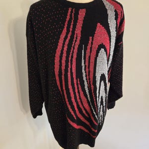 May include: Black sweater with a red, white, and black abstract design. The sweater has a crew neck and long sleeves. The design is a wave-like pattern on the front and a dotted pattern on the sleeves.
