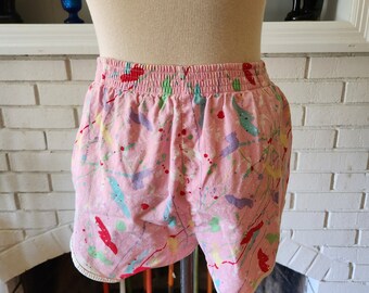 May include: Pink shorts with a paint splatter design. The shorts have a white elastic waistband and a white trim around the leg openings.