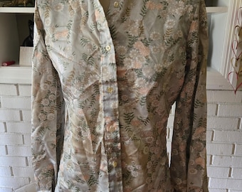 Puede incluir: Una camisa vintage abotonada con cuello alto, exhibida en un maniquí. La camisa presenta un estampado floral en tonos grises, melocotón y verde. Las mangas largas tienen puños con volantes y la camisa está abotonada por delante.