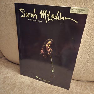 May include: A black music book titled "Sarah McLachlan" with the text "PIANO VOCAL GUITAR" and "INCLUDES SPECIAL GUITAR SECTION". The cover features a dark image of Sarah McLachlan singing.