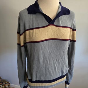 May include: A light blue long-sleeve sweater with a navy blue collar and accents. The sweater features a horizontal band of tan with a diamond pattern, bordered by burgundy stripes. The cuffs and hem have navy, tan, and light blue stripes.