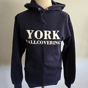 May include: A navy blue hooded sweatshirt with the white text "YORK WALLCOVERING" across the chest. The sweatshirt has a front pocket and a drawstring hood. The garment is displayed on a mannequin.