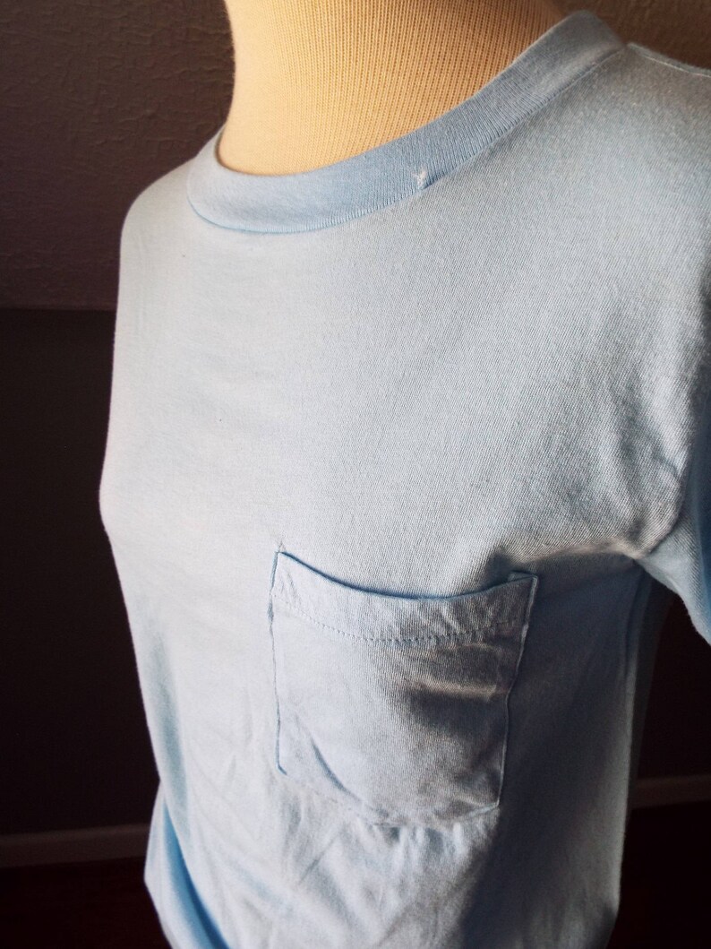 May include: A light blue t-shirt with a single pocket on the left side.