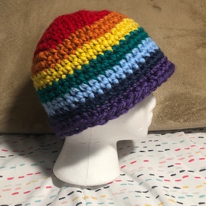 May include: A rainbow crocheted beanie hat with a purple brim. The hat is made with different coloured yarn, including red, orange, yellow, green, blue, and purple.