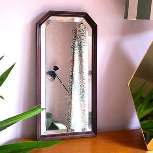 May include: A rectangular, dark brown framed mirror with an octagonal shape. The mirror reflects a patterned curtain, a black lamp, and a glimpse of a room. The mirror is placed on a wooden surface.