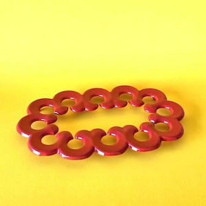 May include: A red, circular trivet with a repeating, stylized design. The trivet is made of a glossy material and sits against a bright yellow background. The design is composed of connected, abstract shapes.