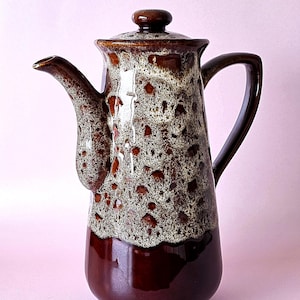 May include: A ceramic coffee pot with a lid and handle. The pot has a speckled brown and white pattern on the upper section, leading to a solid, glossy dark brown base. The spout is long and curved, typical of a coffee pot.