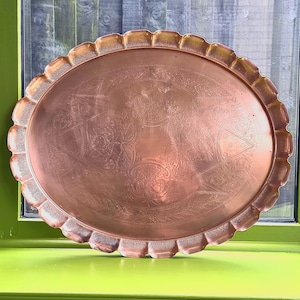 May include: An oval copper tray with a scalloped edge. The tray features an embossed floral and geometric design in the centre. The copper has a warm, reddish-brown hue, and the tray is displayed against a green background.