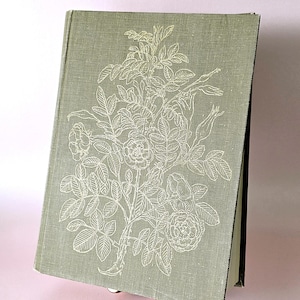 May include: A vintage book with a sage green cover and a white floral design. The cover has a textured appearance, and the book is slightly open, revealing black-edged pages. The floral design includes roses and leaves.