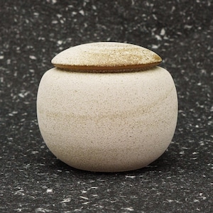 以下が含まれることがあります： 蓋付きの小さな丸い白い陶器の壺。壺は斑点のある質感と薄い茶色の縁をしています。