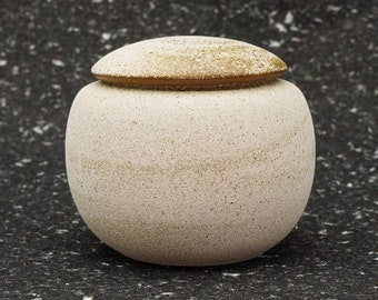 May include: A small, round, white ceramic jar with a lid. The jar has a speckled texture and a light brown rim.