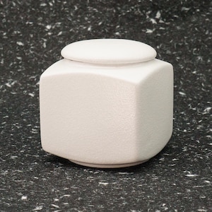 Op de afbeelding: Een vierkante urn van wit keramiek met een afgerond deksel. De urn heeft een getextureerd oppervlak.