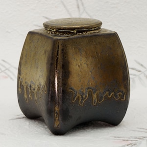 May include: A square, ceramic inkwell with a lid. The inkwell is a dark bronze color with a gold-colored top and dripping design around the base. The inkwell has a unique, curved shape.
