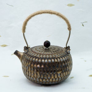 Peut inclure: Une théière en céramique dorée et brune avec une surface texturée et à motifs. La théière a une poignée en corde et un bouton brun foncé sur le couvercle.