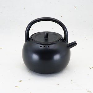 Könnte beinhalten: Eine mattschwarze Keramik-Teekanne mit rundem Körper, gebogenem Henkel und kleinem Ausguss. Der Deckel hat einen kleinen Knopf. Die Teekanne hat drei kleine Löcher in der Nähe des Ausgusses. Der Hintergrund ist hell mit Flecken.