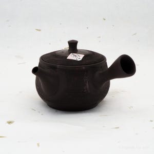 Può includere: Una teiera in ceramica marrone scuro con coperchio e manico laterale. La teiera ha una superficie strutturata e un piccolo beccuccio sul lato opposto al manico. Una piccola etichetta è apposta sul coperchio.