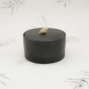 Puede incluir: Un recipiente de cerámica negra con tapa. La tapa tiene ranuras circulares concéntricas y un pequeño asa de cuerda de color beige. El recipiente tiene forma cilíndrica con una banda decorativa cerca de la base.