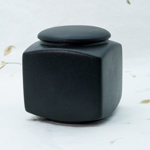 Puede incluir: Un recipiente cuadrado de cerámica negra con tapa. El recipiente tiene una superficie texturizada.