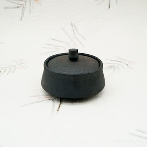 Può includere: Un piccolo vaso in ceramica grigio scuro con coperchio. Il vaso ha un corpo arrotondato e un coperchio piatto e circolare con un piccolo pomello sulla parte superiore. La superficie del vaso e del coperchio appare strutturata. Sfondo bianco con motivo floreale.