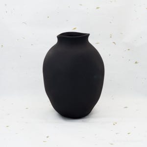 Puede incluir: Un jarrón de cerámica negro mate con un cuerpo redondeado y un borde ligeramente ensanchado e irregular. El jarrón tiene una superficie lisa y no reflectante y un diseño simple y elegante. El jarrón es adecuado para exhibir flores o como objeto decorativo.