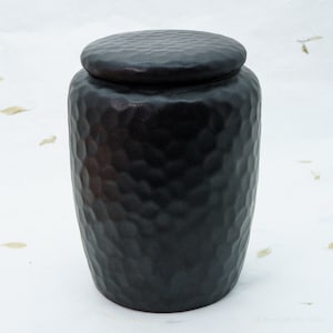 Peut inclure: Un pot en céramique noire avec une surface texturée et un couvercle. Le pot est rond et a une base légèrement plus large que le haut.