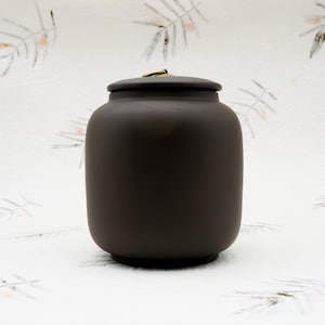 Op de afbeelding: Een donkerbruine keramische urn met een deksel en een kleine goudkleurige handgreep. De urn heeft een afgeronde vorm en staat op een platte basis. De achtergrond is een wit oppervlak met een subtiel bloemmotief.