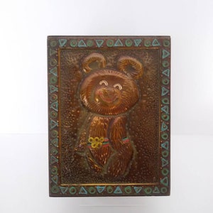 May include: A rectangular, bronze-colored metal plaque featuring a smiling bear with the Olympic rings. The bear is embossed with details, and the border has a decorative pattern of triangles and circles in teal and gold.