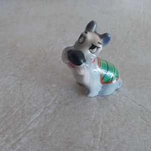 May include: A small ceramic figurine of a gray and white dog wearing a green and orange vest. The dog has black eyes and a black nose.