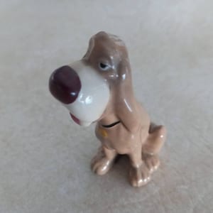 May include: A ceramic figurine of a brown and white bloodhound dog with a brown nose and a yellow collar. The dog is sitting with its head tilted to the side.