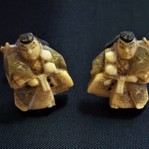 May include: Two small ivory carvings of Japanese figures in traditional clothing. The figures are holding fans and are intricately detailed.